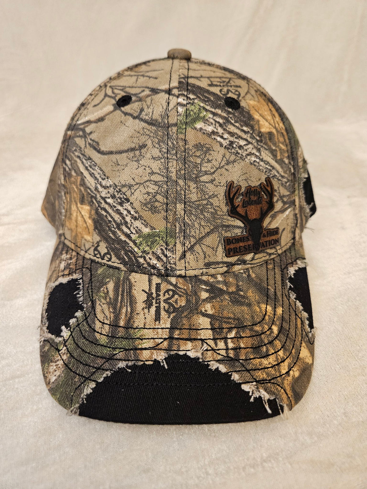 Camo hat with black distressed detailing and engraved leather Bones & Hide Preservation patch, front
